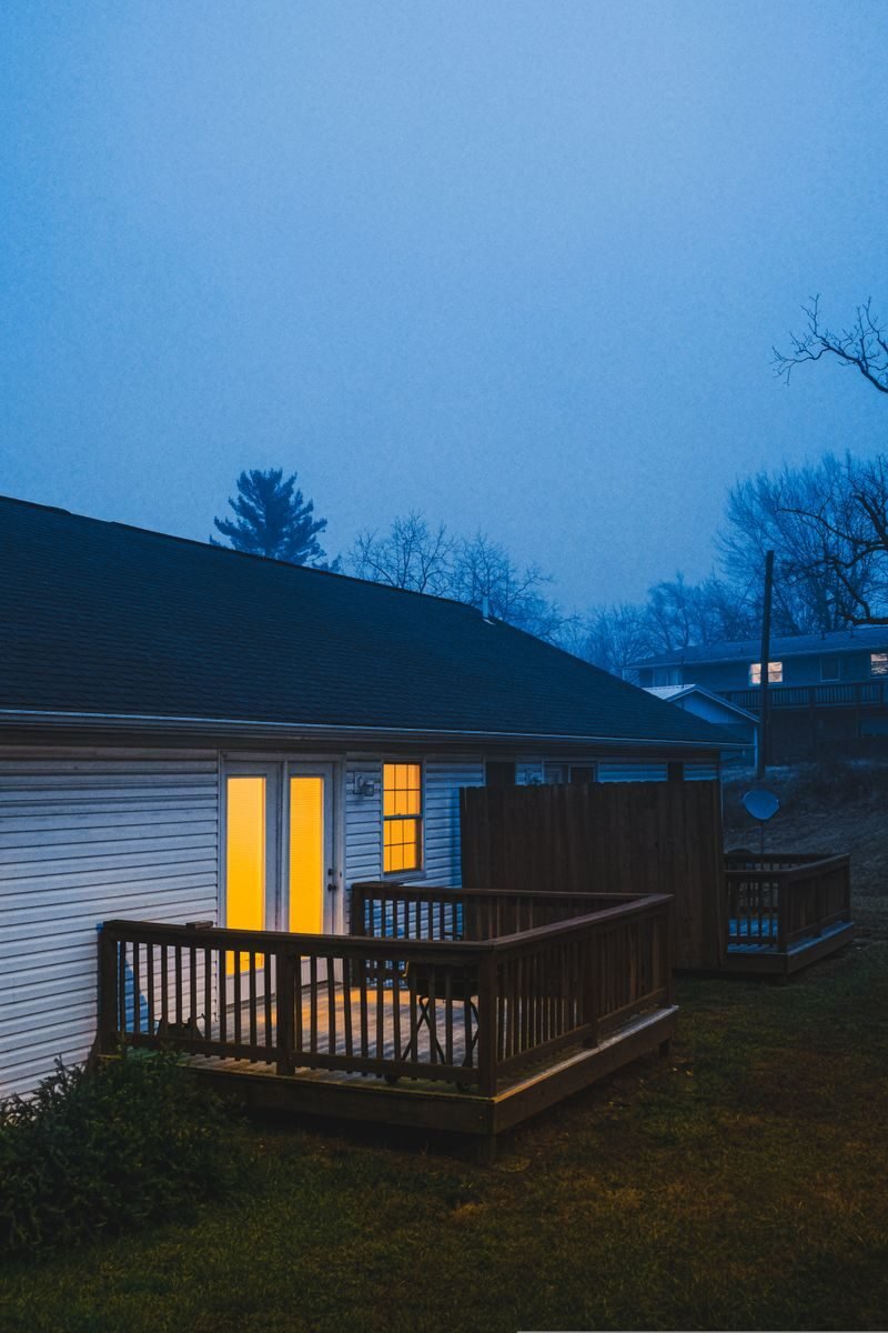 A suburban house with a warmly lit porch on a misty evening outdoors.